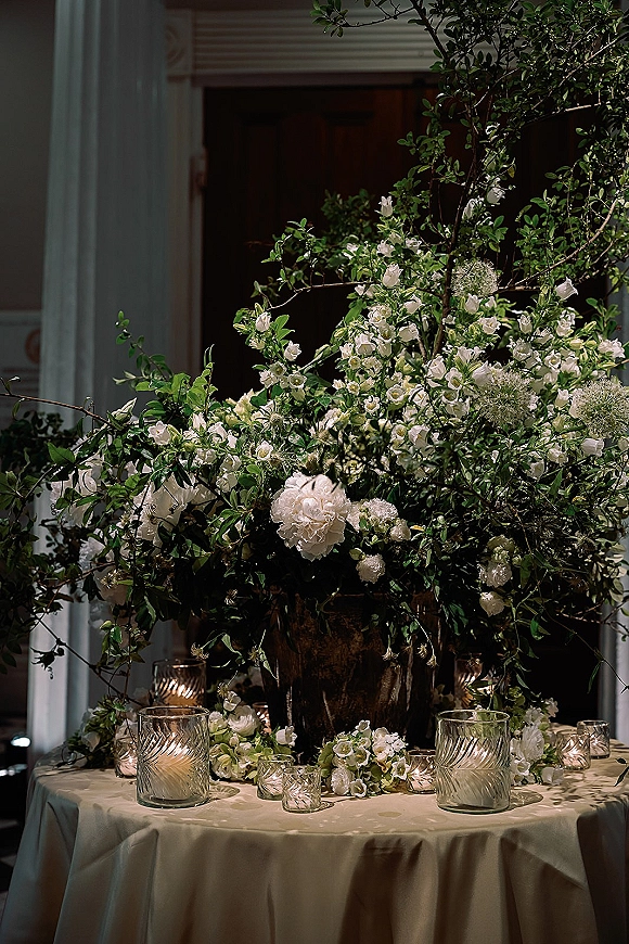 Reception tablescape with a wedding floral centerpiece of white florals and greenery, set with candle votives in glass holders by a doorway