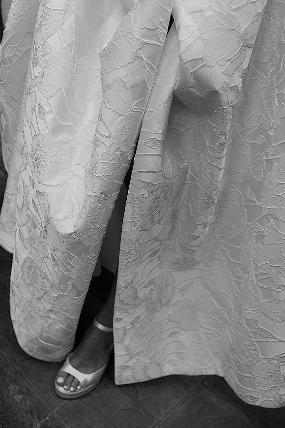 Wedding dress detail showing floral jacquard wedding dress fabric with a high slit, open-toe heels, and hem on a stone floor