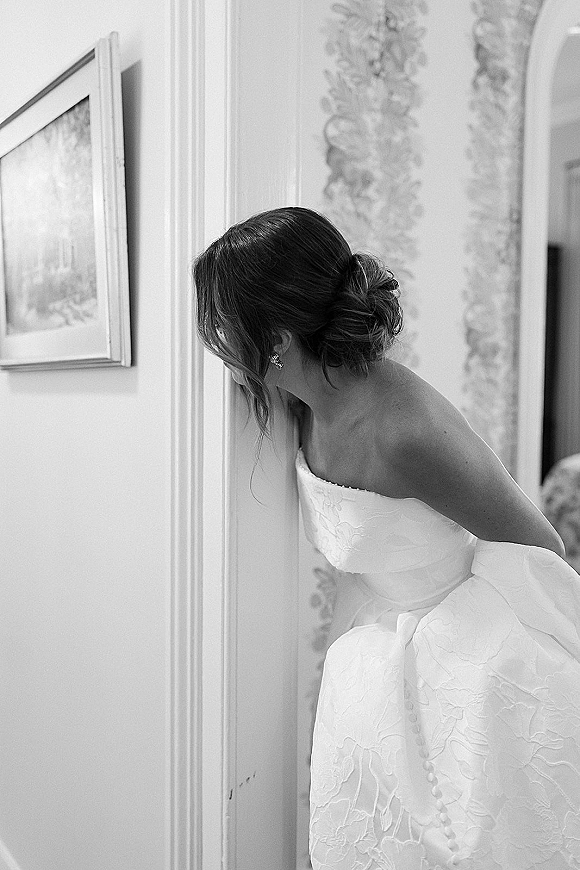 Bridal portrait of bride peeking around a doorway in a hallway, strapless textured gown with low bun, loose tendril, and drop earrings