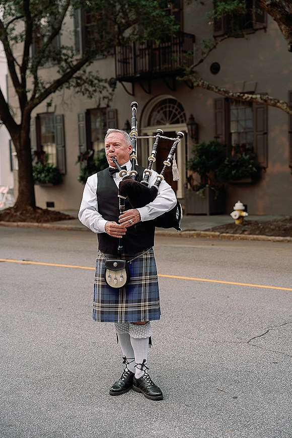 Wedding bagpiper playing in tartan kilt on a tree-lined street, passing townhouse balconies and shutters in natural daylight