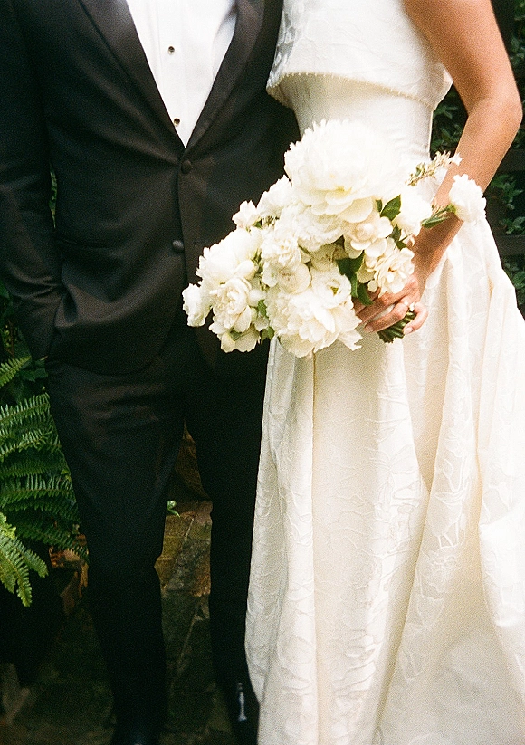 Couple portrait of bride and groom holding a white bridal bouquet on a stone garden path, her textured gown beside his black tuxedo