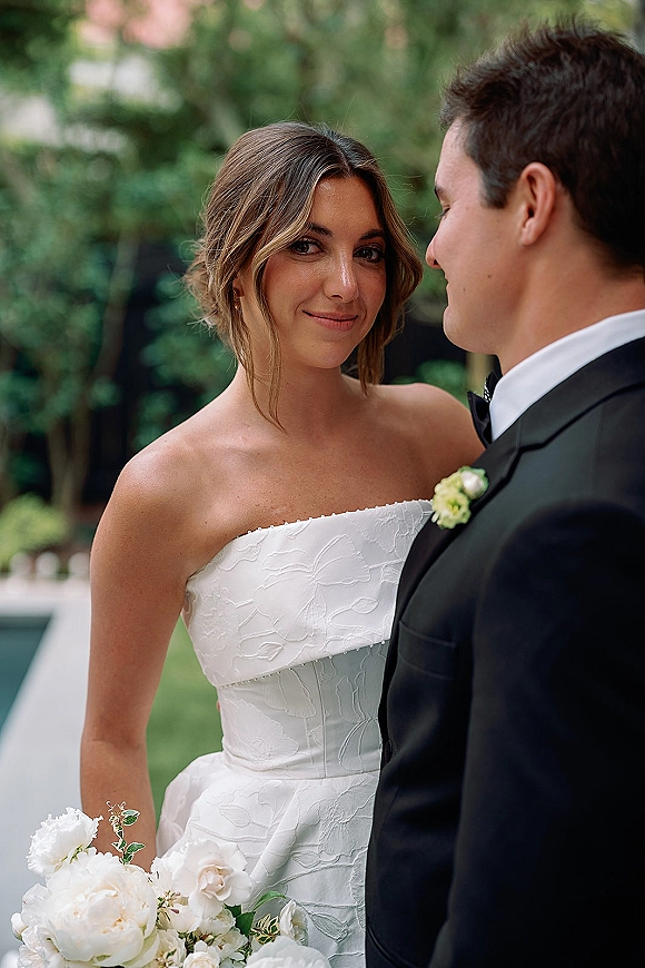 Couple portrait of bride and groom portrait embracing, bride in strapless dress holding a white peony bouquet against blurred garden greenery