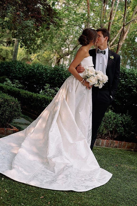 Wedding kiss portrait of bride and groom kissing, her strapless gown and white bouquet with greenery in a tree-lined garden with hedges