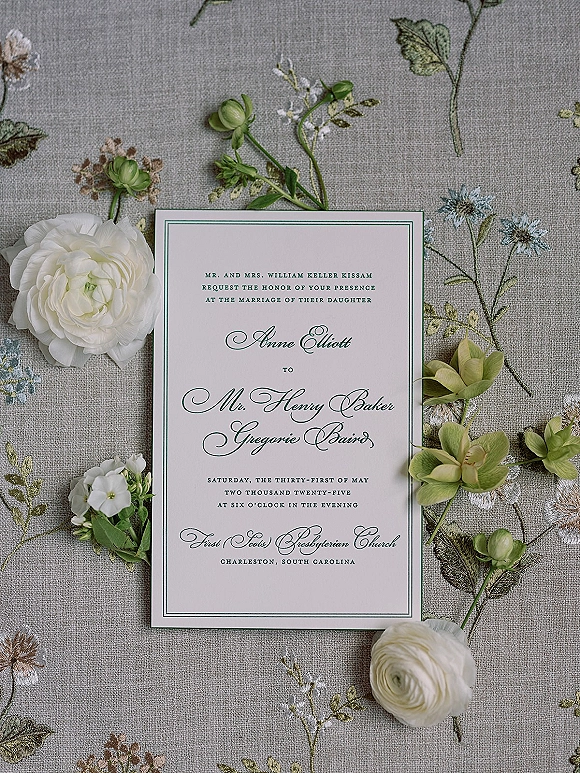 Wedding invitation with letterpress design, green border and calligraphy script beside white ranunculus and orchid blooms on embroidered linen
