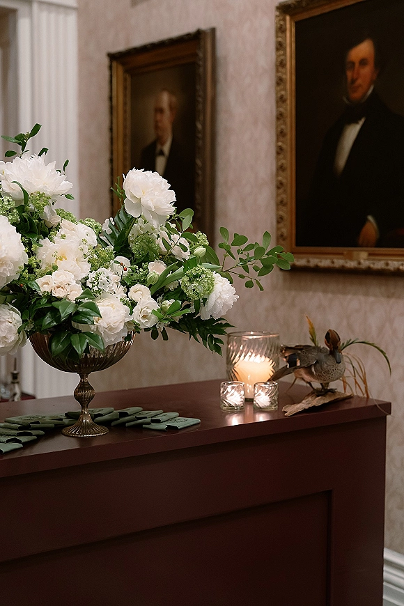 Wedding welcome table decor with white flowers and greenery in a metal compote vase, candles and velvet ribbon escort cards, portraits behind
