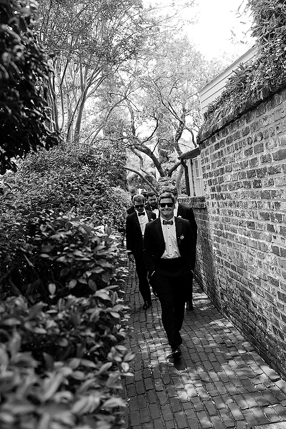 Groomsmen portrait of the groom with groomsmen in tuxedos and bow ties, wearing sunglasses on a brick walkway by hedges and trees
