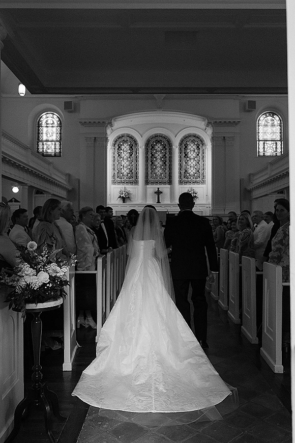 Wedding processional of bride walking down aisle with cathedral veil and long train, guests in pews beneath stained glass windows