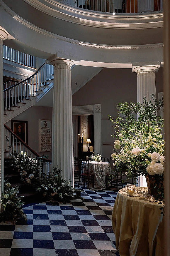 Wedding venue decor with white floral arrangements and greenery on a round table with votive candles in a grand staircase foyer