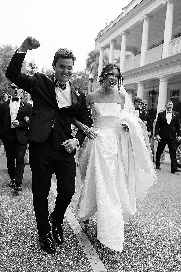 Wedding recessional just married exit as bride in strapless dress and veil and groom in tux cheer past guests by columned facade and trees