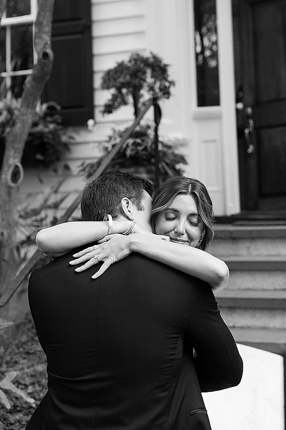 First look moment as bride hugs groom from behind, eyes closed, engagement ring visible on porch steps outside a house with plants