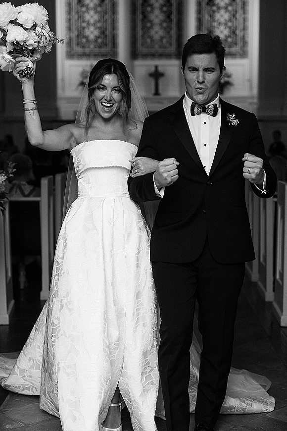 Wedding recessional as just married walk down a church aisle, bride lifting bouquet and veil beside groom in tuxedo, stained glass behind