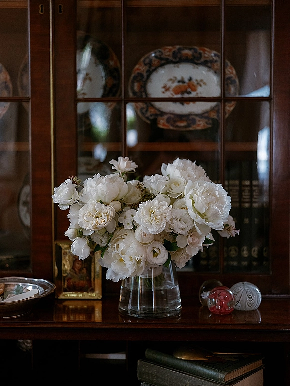 Wedding flowers in a white peony arrangement with white roses and greenery in a clear glass vase on a wood cabinet before a glass-front hutch