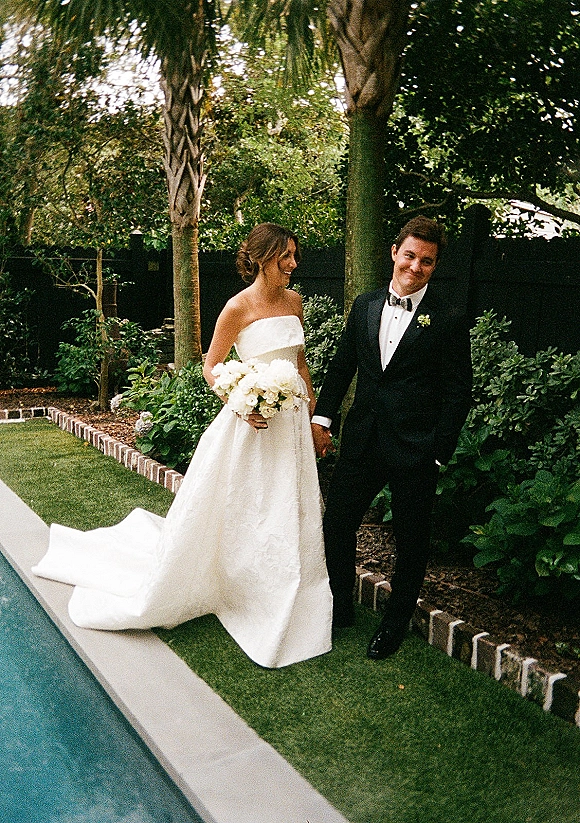 Couple portrait of bride in a strapless gown holding a white rose bouquet with groom in tuxedo by a pool and palm-lined garden lawn