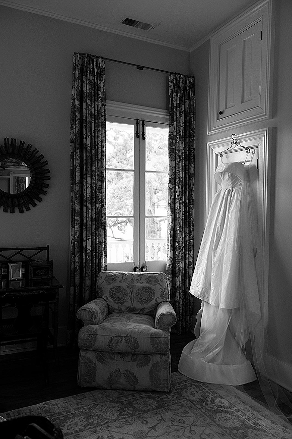 Wedding dress hanging from a hanger, strapless lace gown with long train by French doors in a bedroom with soft window light