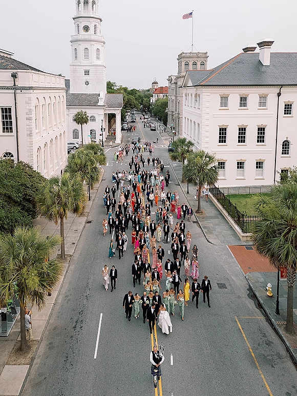 Wedding procession with bride and groom leading a lively group down a city street, passing historic buildings and a church steeple, American flag waving