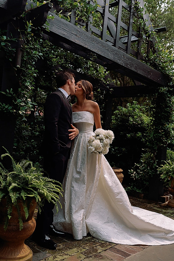 Wedding kiss portrait of bride and groom kissing, her strapless gown with long train and rose bouquet by a garden staircase on brick patio