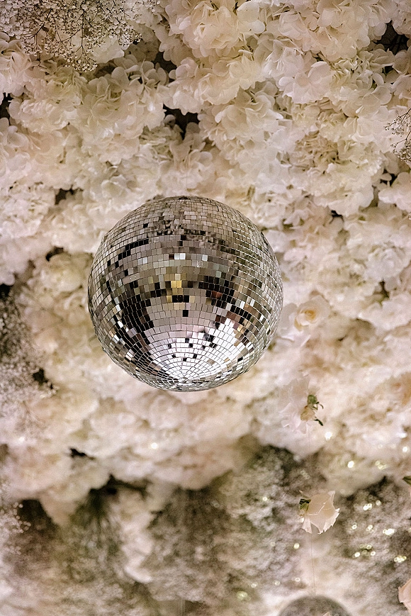 Disco ball decor with a mirror disco ball hanging among white hydrangeas and baby’s breath under twinkling string lights on a floral ceiling