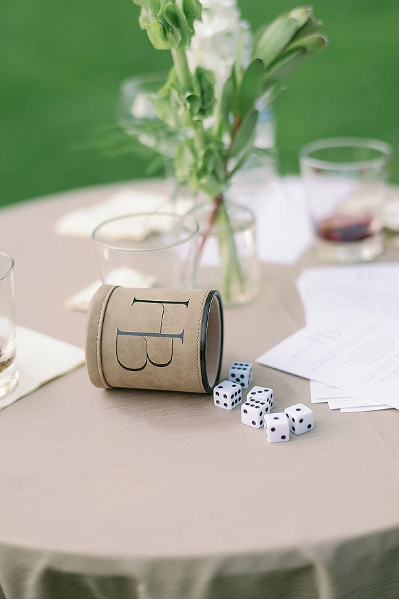 Wedding reception table with place cards, printed stationery, cocktail glasses, white floral centerpiece and dice on a green lawn outdoors
