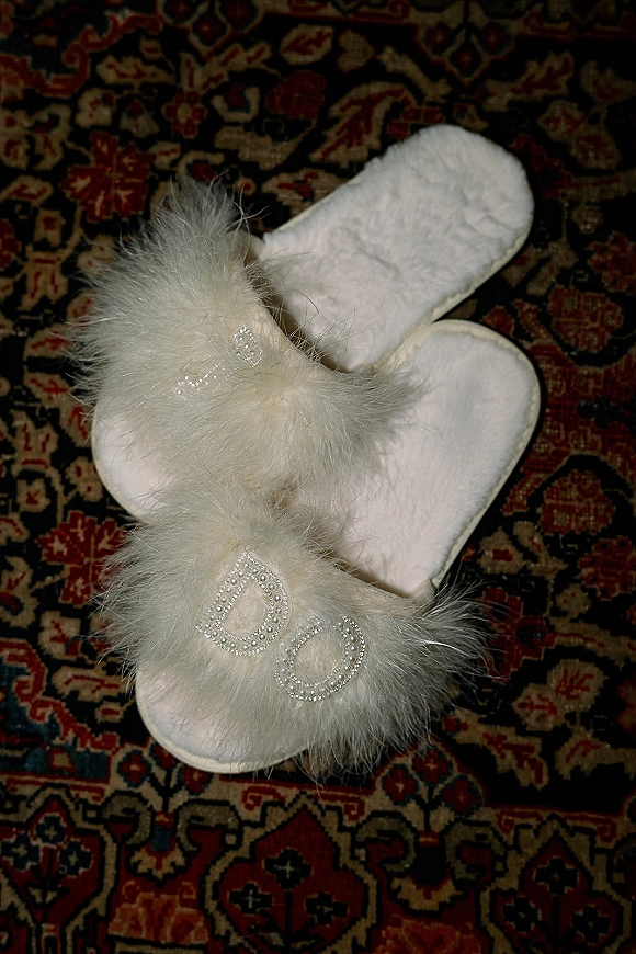 Bridal slippers with feather trim and rhinestone lettering in a bride slippers flatlay on a patterned rug, cozy wedding day detail