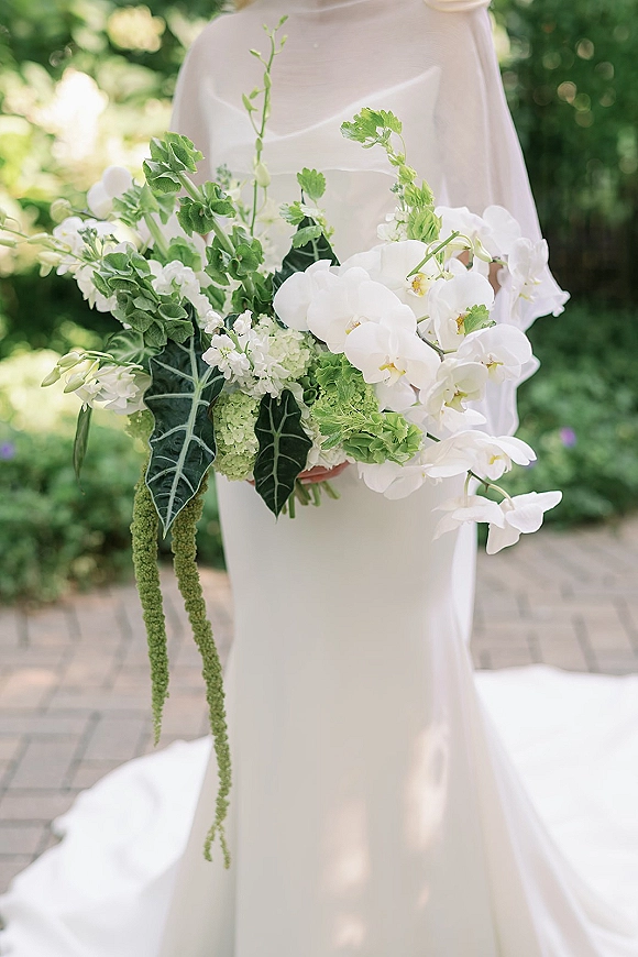 Bridal bouquet with white orchid blooms and greenery, cascading with trailing amaranthus against a strapless satin dress in garden light