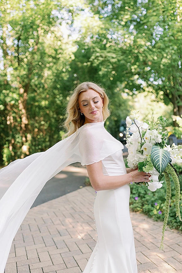 Bridal portrait of a bride with veil blowing behind her, holding a white orchid bouquet on a sunlit, tree-lined path with bokeh greenery