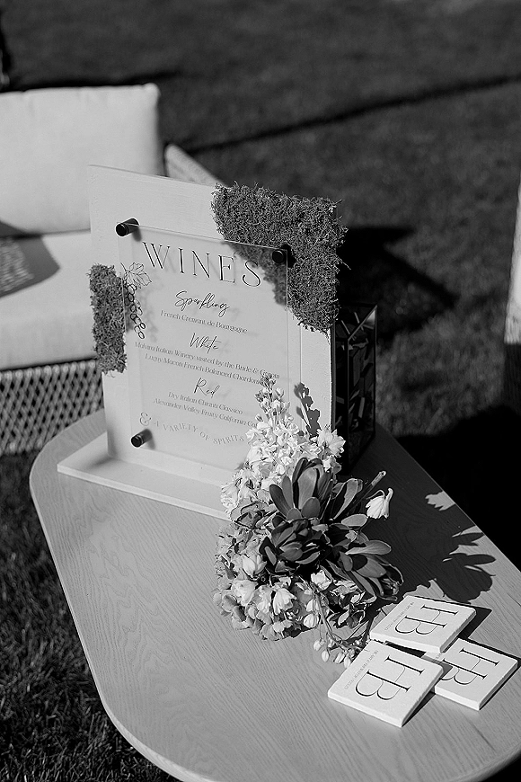 Wedding bar menu acrylic sign with standoff hardware, florals, and moss on a wooden table beside cocktail napkins on a grassy lawn