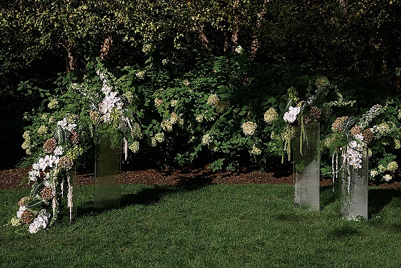 Ceremony floral pillars on clear acrylic pedestals with white orchids and hydrangeas, hanging amaranthus and greenery on a garden lawn