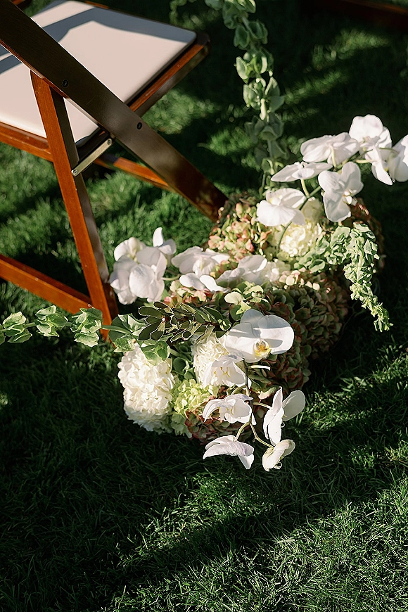 Aisle floral arrangement of white orchids and hydrangeas with eucalyptus draped on a wooden folding chair on a sunlit grass lawn