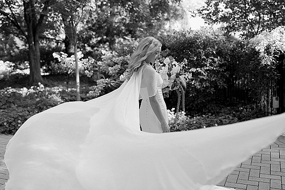 Bridal portrait in black and white of a bride looking down, holding a bouquet, with long train and veil on a garden walkway