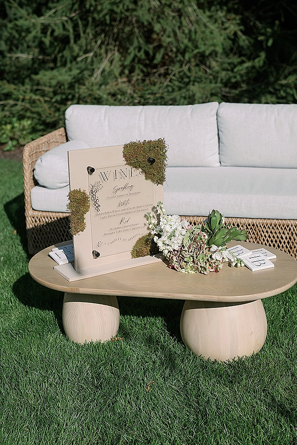 Wedding bar sign on an acrylic stand with moss and floral accents, wine list typography on a wooden table in a garden lounge setup