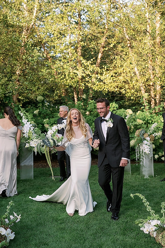 Wedding recessional as bride and groom walk hand in hand down a garden aisle, bride laughing with orchid bouquet, guests behind