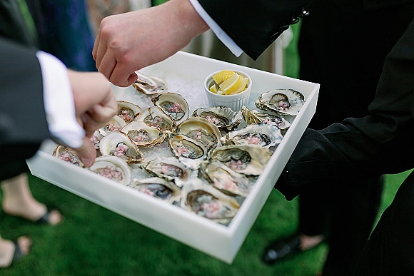 Oyster bar serving tray with oysters on ice and lemon wedges, held by suit sleeves during outdoor wedding cocktail hour on grass lawn