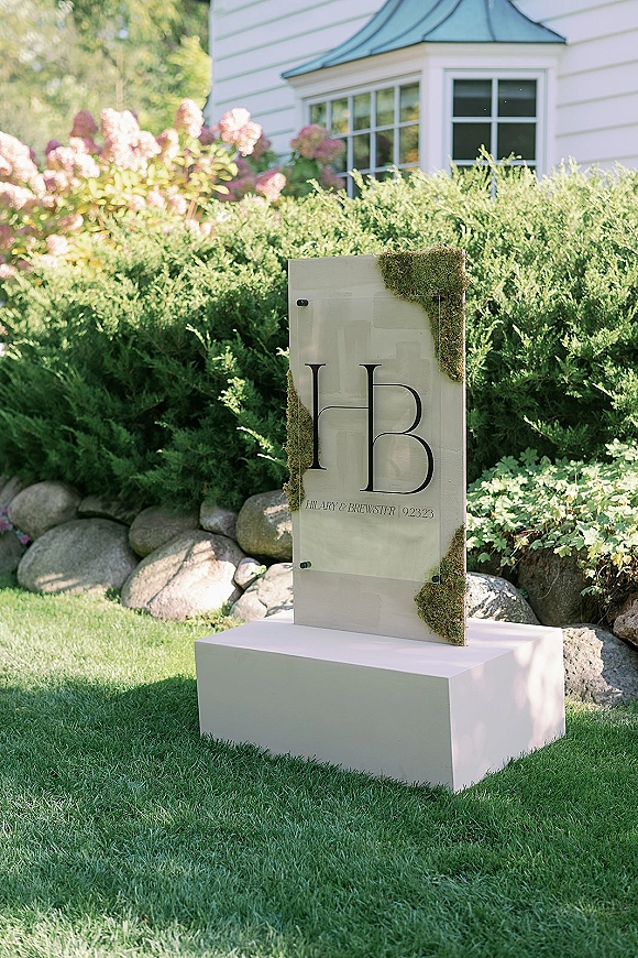 Wedding welcome sign on a clear acrylic panel with monogram lettering and moss accent, mounted on a pedestal in a garden lawn near a house