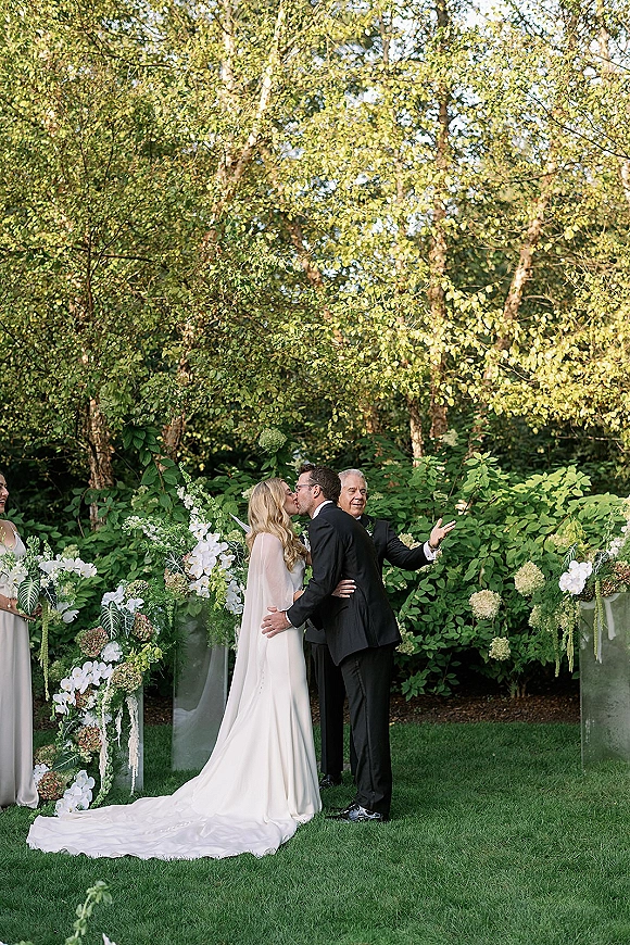 Ceremony kiss at an outdoor wedding ceremony as bride and groom kiss in veil and tux before white florals on glass pedestals.