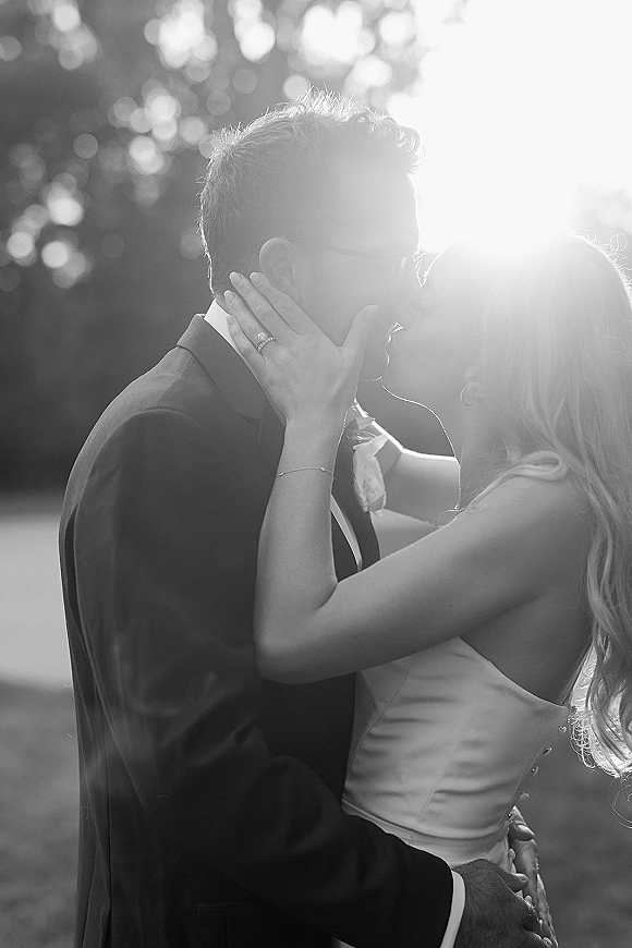 Wedding kiss portrait of bride and groom kissing, her hand on his face showing rings, with sun flare through trees behind them