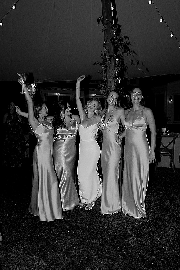 Bridesmaid group photo with bride and bridesmaids in satin dresses raising drinks under string lights in a tented outdoor night reception