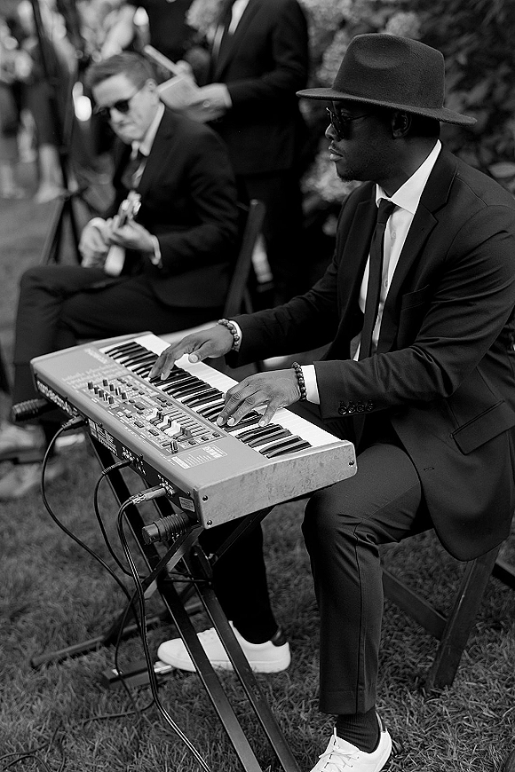 Wedding ceremony musicians, a keyboard player in suit and fedora with sunglasses and a guitarist performing outdoors before seated guests on grass