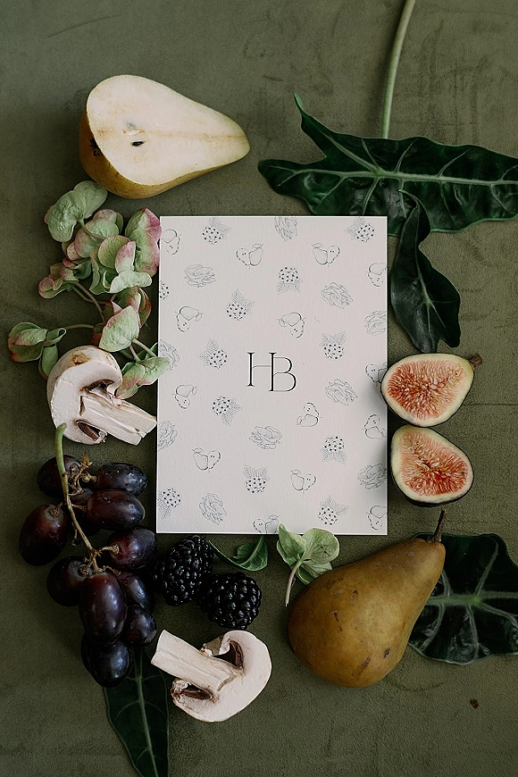 Invitation suite wedding invitation flat lay with monogram card on green fabric, styled with pears, figs, grapes, mushrooms, and greenery