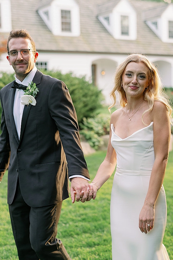 Couple portrait of bride and groom holding hands, her strapless satin gown and his black tuxedo, walking on a lawn by a white estate building