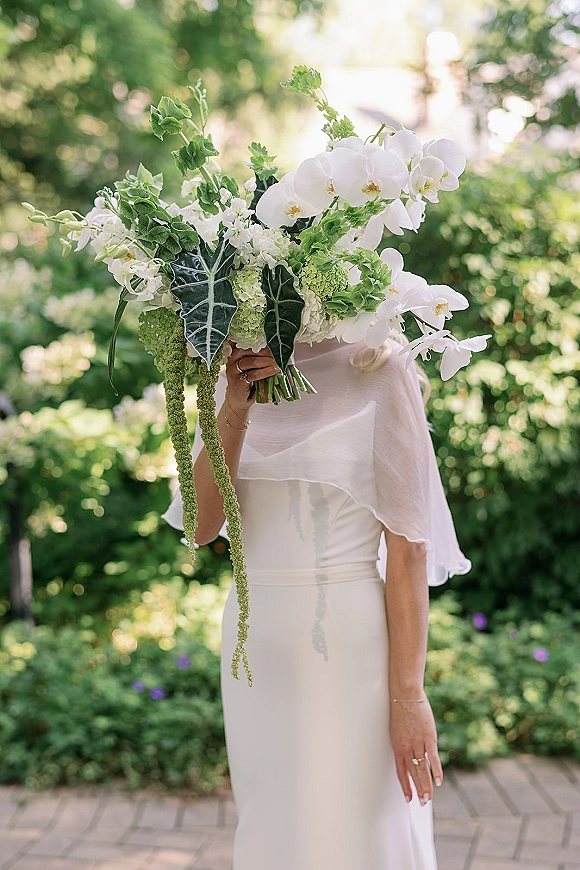 Bridal portrait of a bride holding bouquet over her face, cascading white orchids and greenery in a sunlit garden walkway