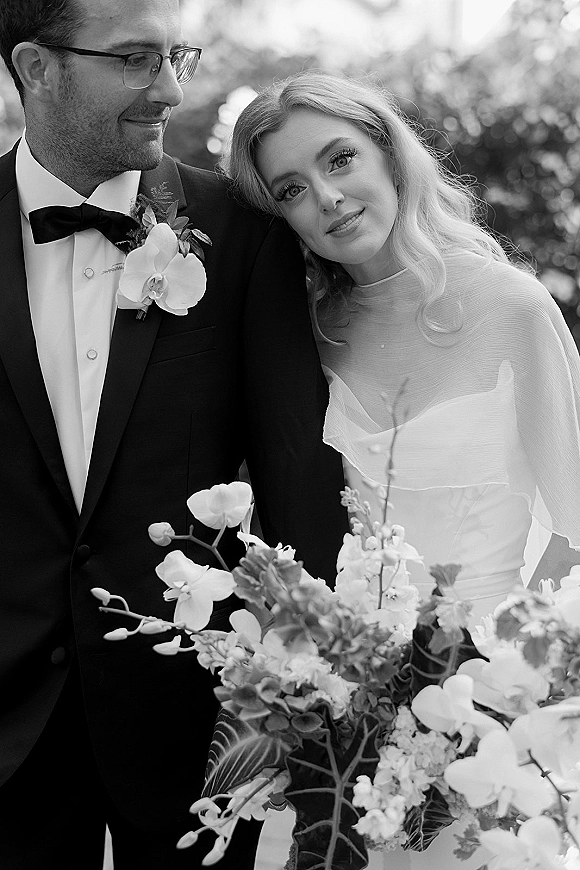 Couple portrait, black and white wedding portrait of bride leaning on groom in tuxedo with orchid boutonniere, outdoor greenery behind
