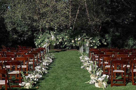 Ceremony setup for an outdoor wedding ceremony with wooden chairs and white aisle flowers on a sunlit garden lawn under trees