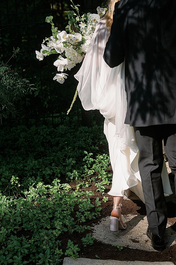 Couple portrait of bride and groom walking from behind, bride holding a white orchid bouquet with veil in dappled garden light