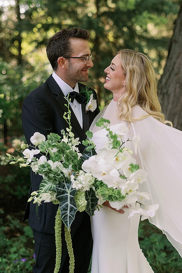 Couple portrait of bride and groom smiling face to face, holding white orchid bouquet in a sunlit garden with soft bokeh trees