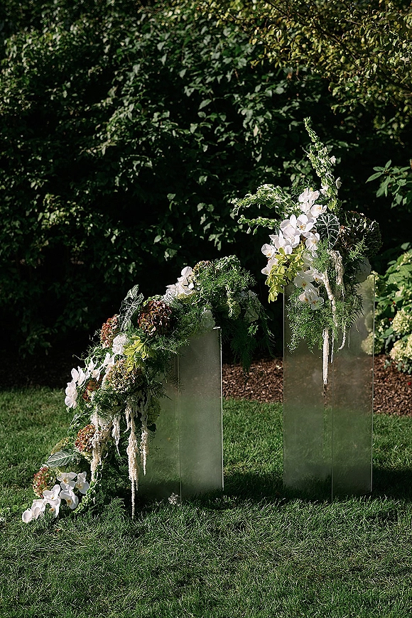 Wedding ceremony backdrop with modern wedding altar on clear acrylic pedestals, white orchids and hanging amaranthus in a garden setting
