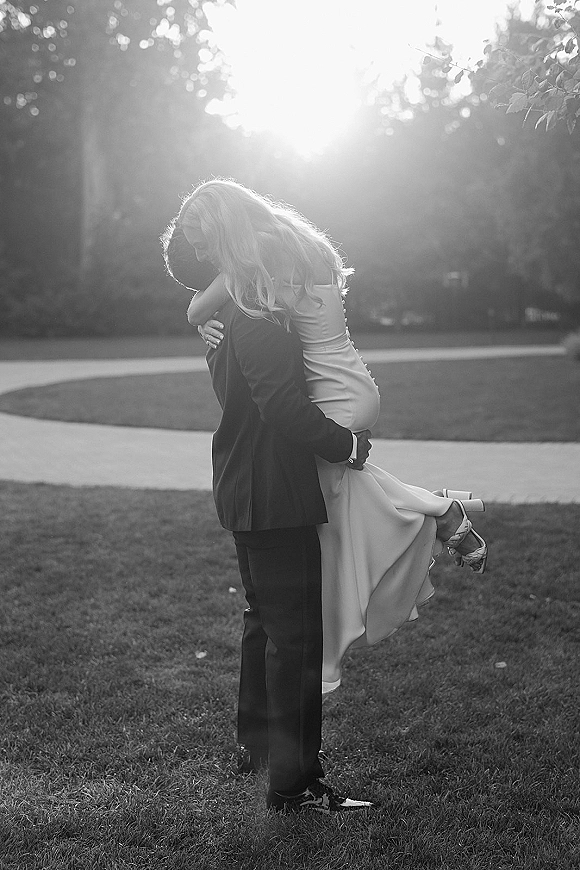 Couple portrait of groom carrying the bride in a wedding lift pose on a tree-lined lawn by a paved path, sunlight behind them