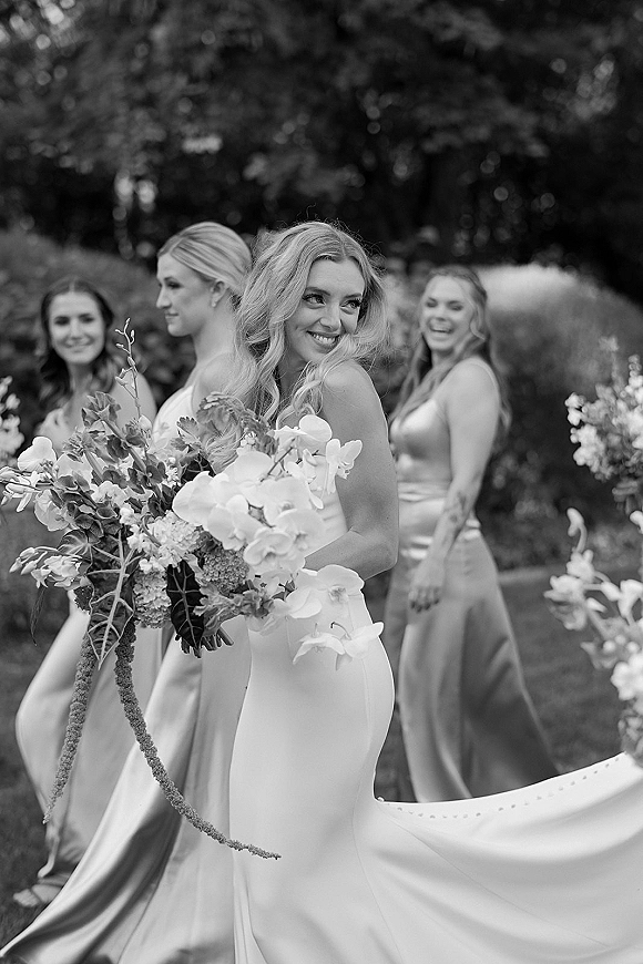 Bride portrait with bouquet, looking over her shoulder in a strapless gown as bridesmaids in satin dresses walk behind in a garden