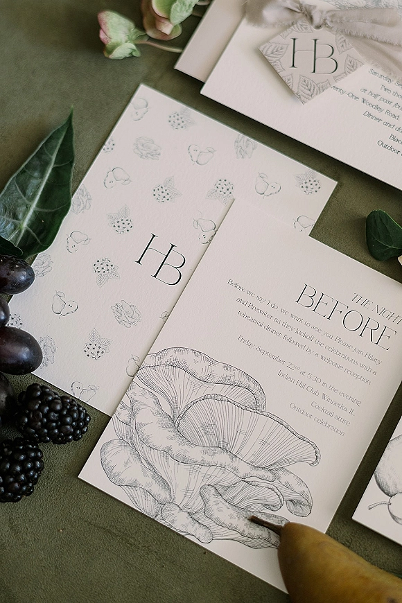 Invitation suite with monogram wedding invitations, patterned envelopes, ribbon, and fruit accents on an olive green fabric flatlay background