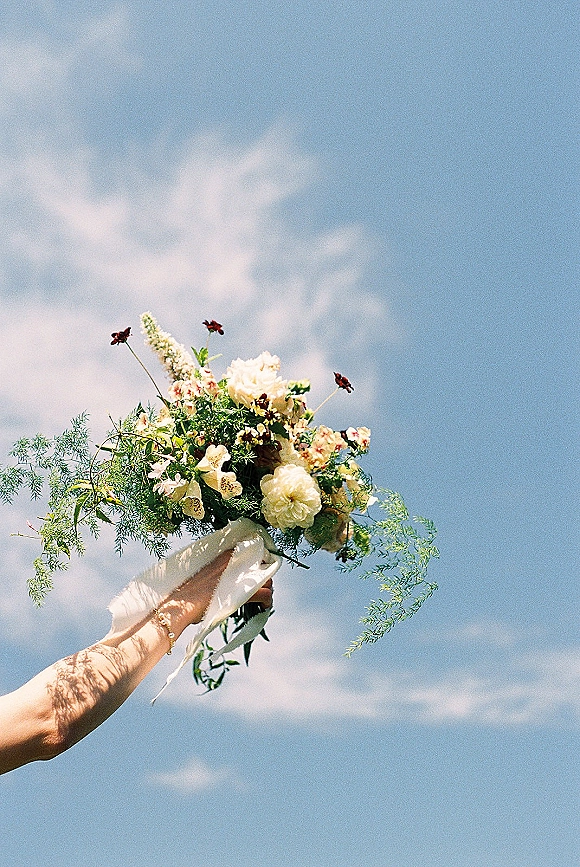 Bridal bouquet of wildflowers and greenery, hand-tied with a white ribbon, held up against a blue sky with clouds