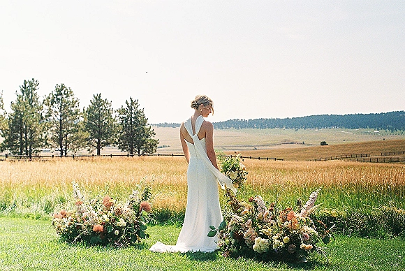 Bridal portrait of a bride holding bouquet in a backless halter wedding dress, looking over her shoulder in a meadow of tall grass